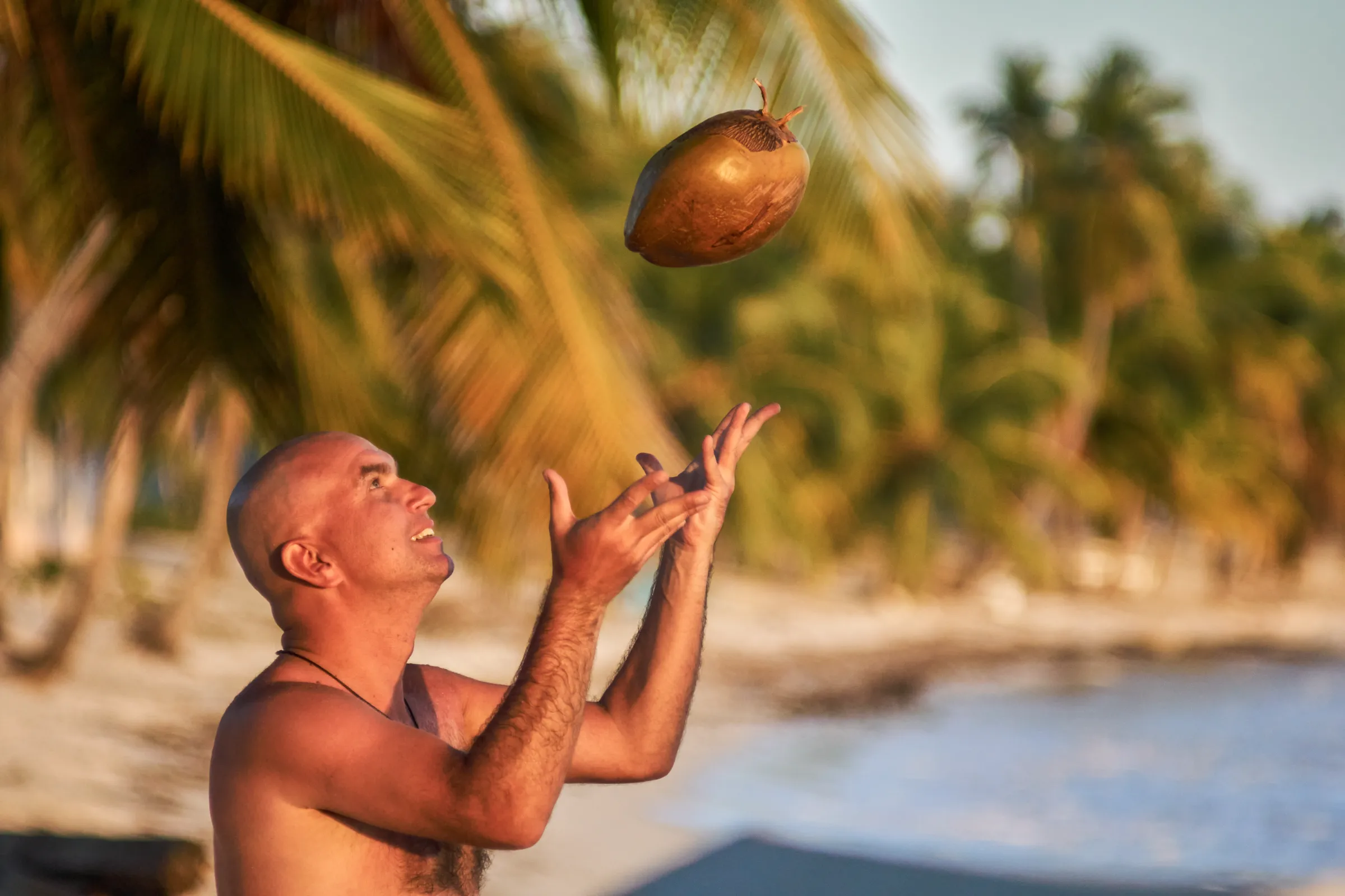 Juego y Luz en la Playa de Santo Domingo Hombre en playa de Santo Domingo lanza coco al aire mientras sonríe, con palmeras y mar de fondo, luz dorada natural de mañana, sesión familiar fotográfica