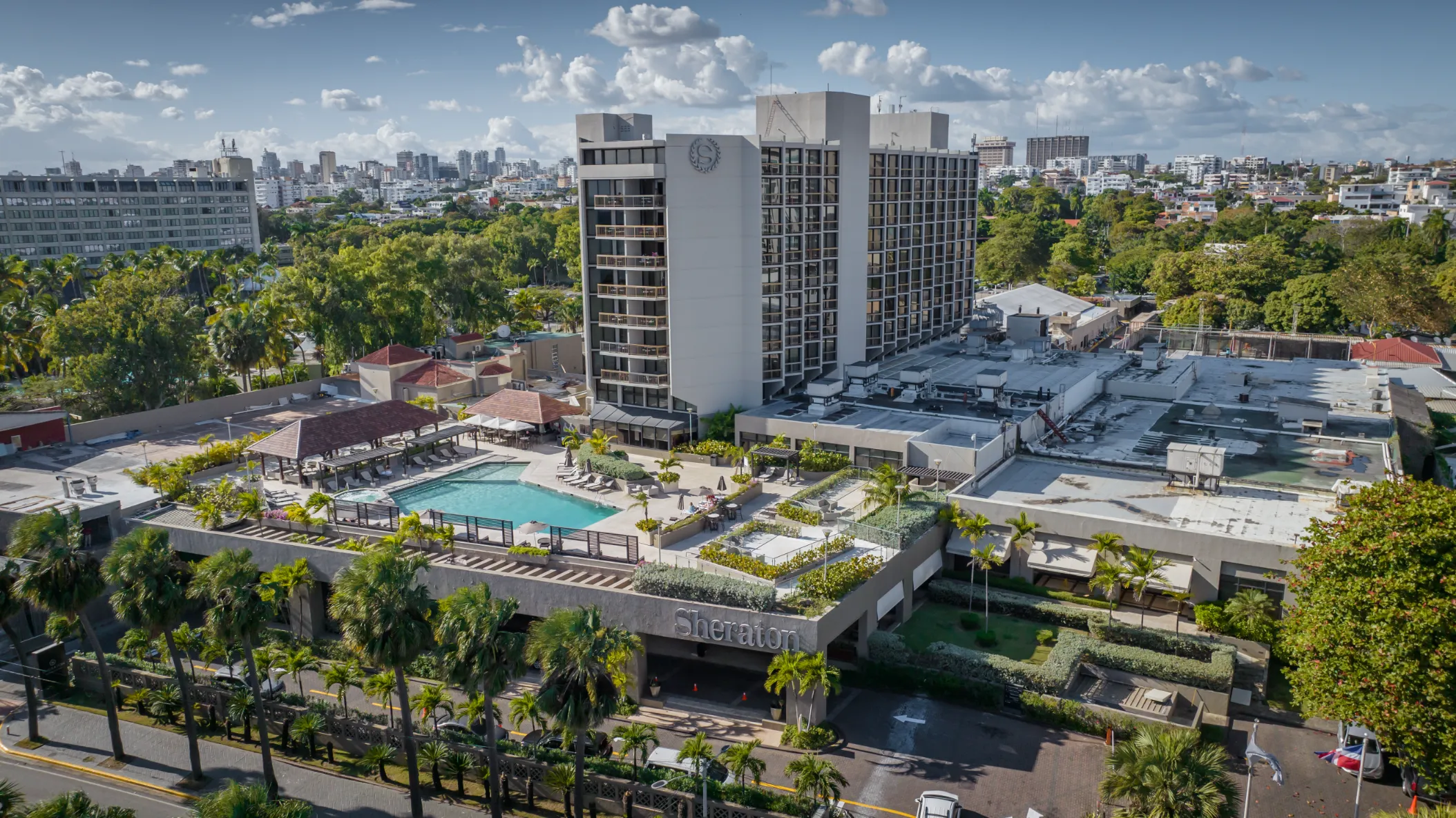 Complejo Residencial y Piscina desde el Cielo Fotografía aérea de complejo residencial en Santo Domingo: piscina turquesa, torre blanca, palmeras y paisaje urbano bajo cielo tropical.