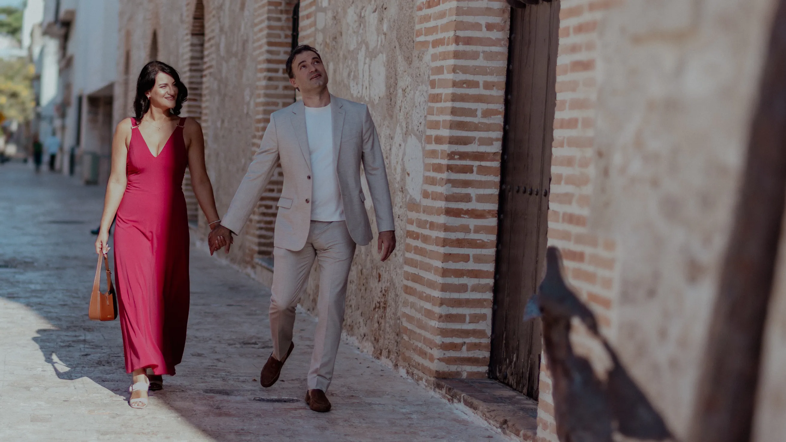 Amor en movimiento por la Zona Colonial Pareja caminando en las calles empedradas de la Zona Colonial de Santo Domingo; ella viste rojo intenso, él traje beige. Luz natural del mediodía