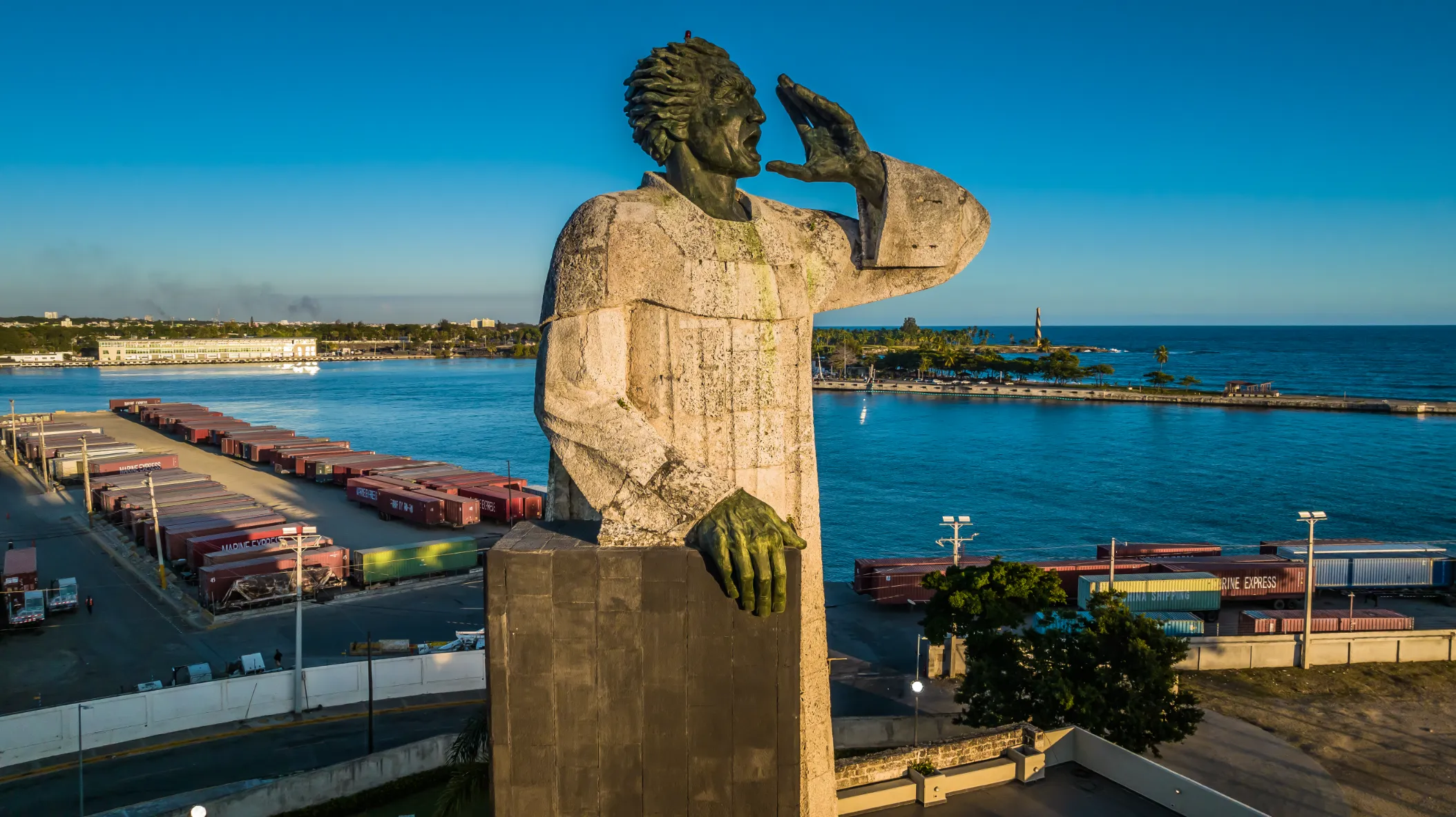 Monumento portuario en luz matinal dorada Estatua de piedra dorada con mano en mejilla frente a puerto de Santo Domingo con contenedores y agua turquesa, fotografía comercial