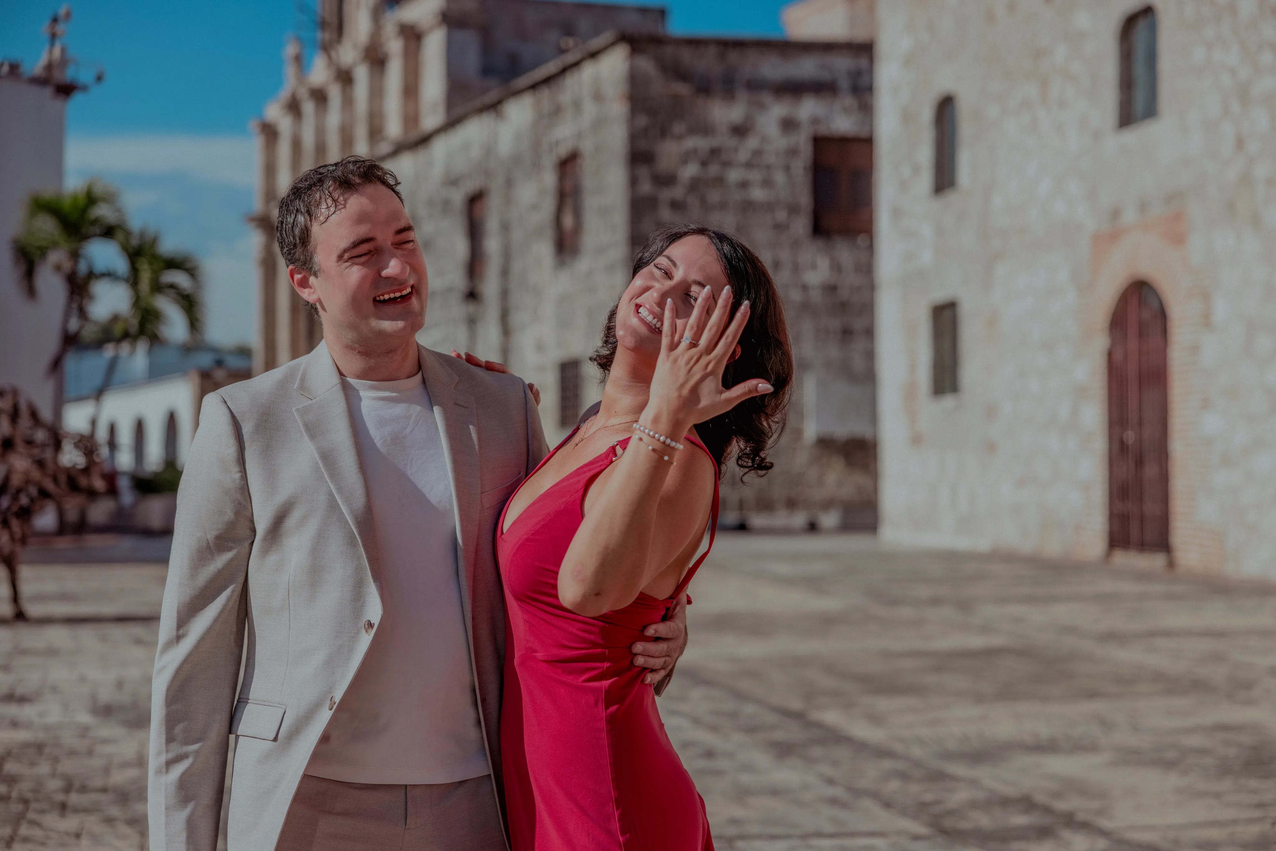 Sesión de Pareja en Santo Domingo Pareja riendo en plaza del casco antiguo de Santo Domingo, ella con vestido rojo neón, él con blazer beige, arquitectura colonial blanca