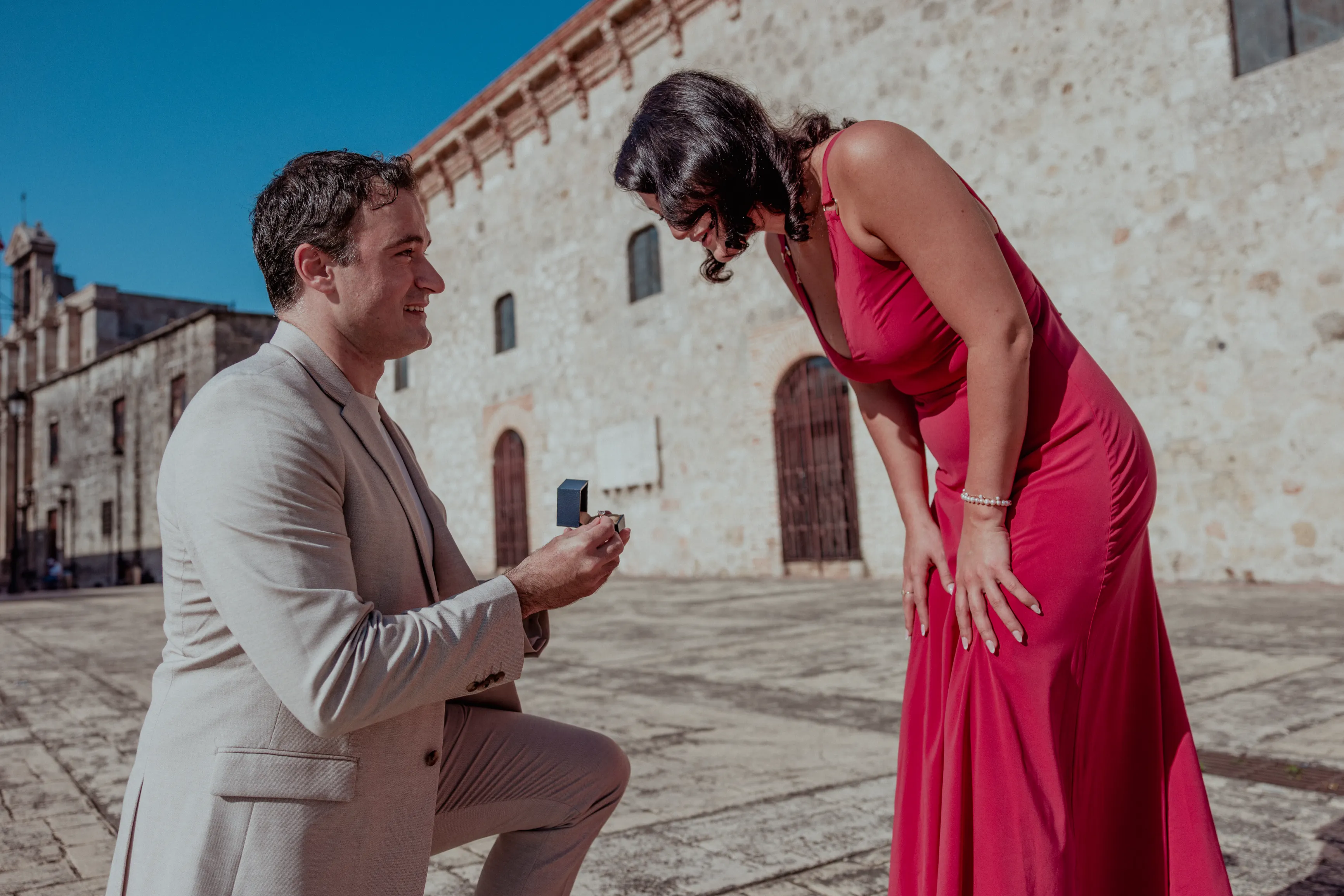 Propuesta de matrimonio bajo el cielo de Santo Domingo Hombre en traje beige propone matrimonio con anillo de diamante a mujer en vestido fucsia en plaza colonial de Santo Domingo