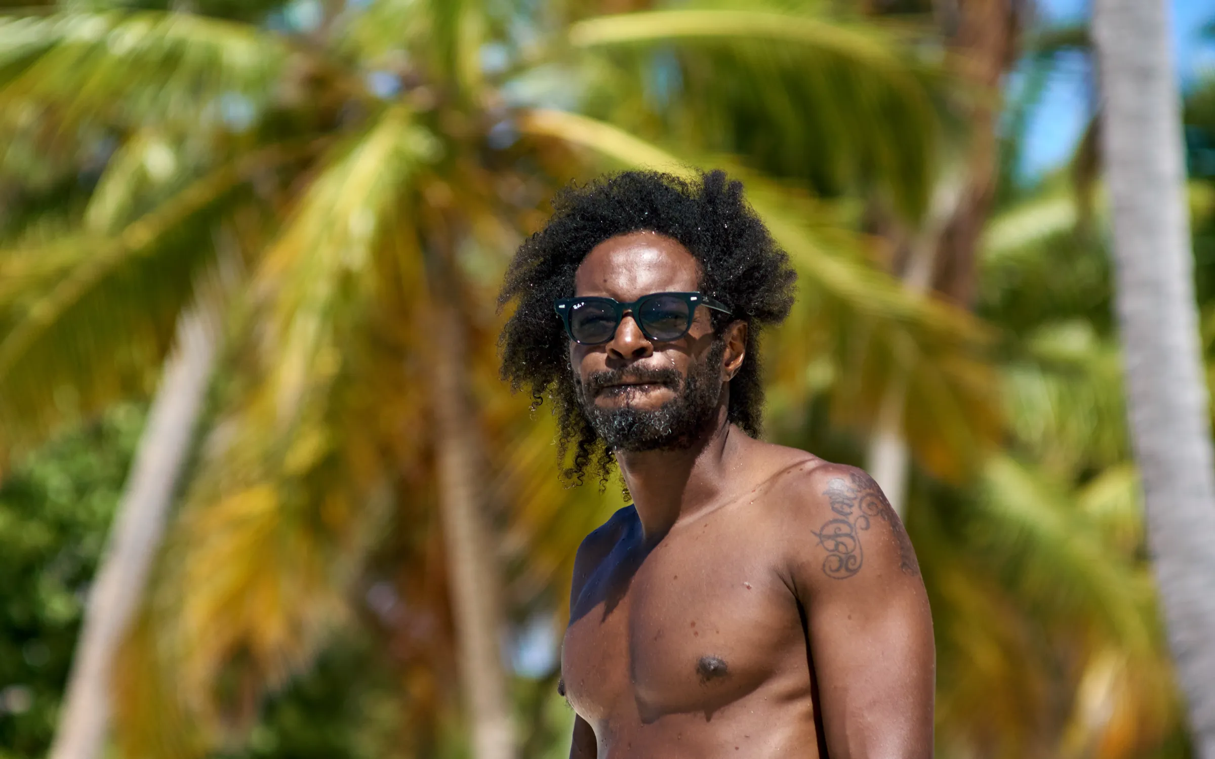 Retrato de Lujo en Playa Tropical Hombre con gafas oscuras posando en playa tropical con vegetación de palmas desenfocadas, sesión de retrato de lujo en Punta Cana, República Dominicana