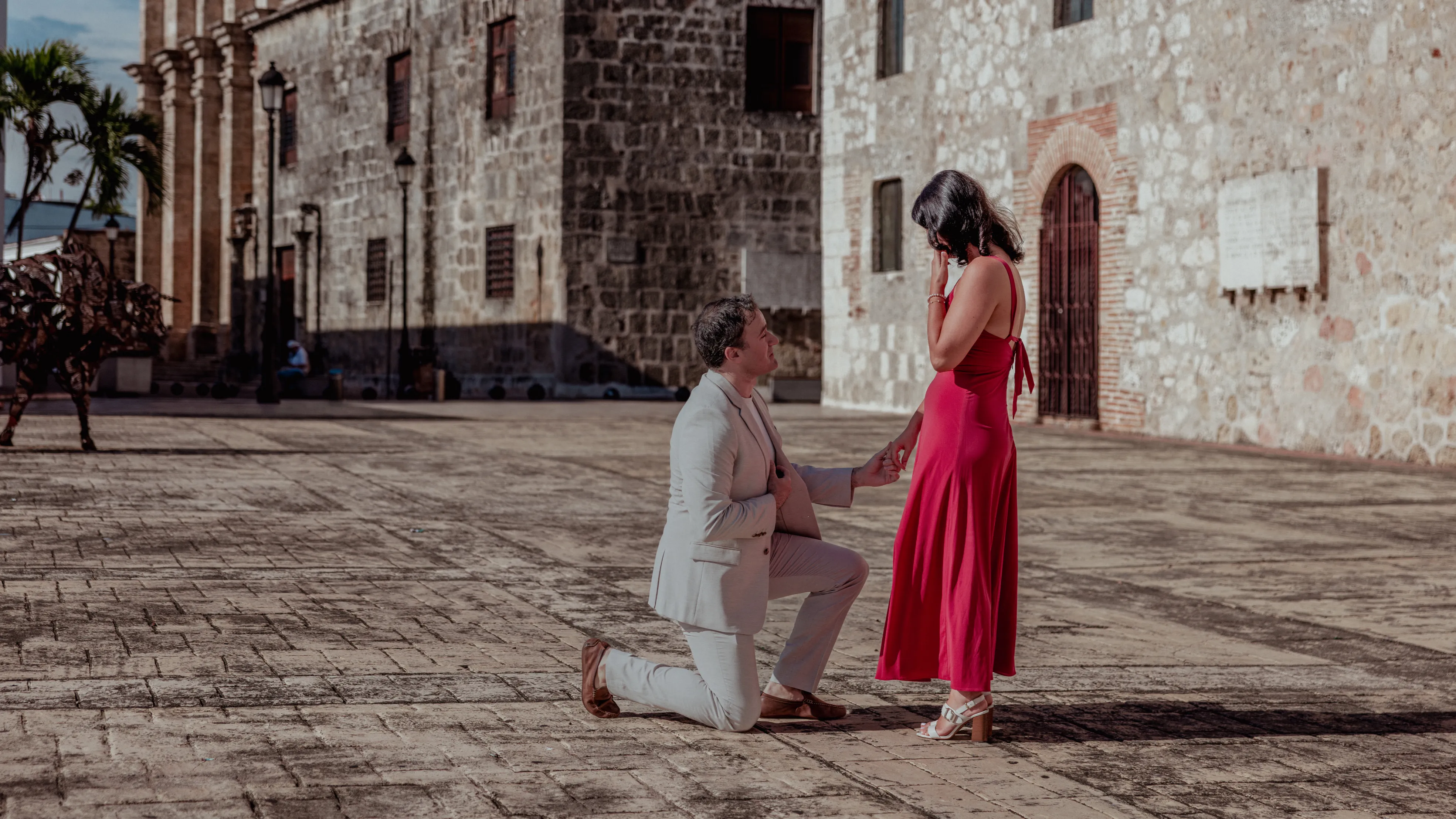 El sí susurrado en la Plaza Colonial Propuesta de matrimonio en Santo Domingo: hombre de rodillas en plaza colonial frente a mujer con vestido rojo, luz lateral de tarde